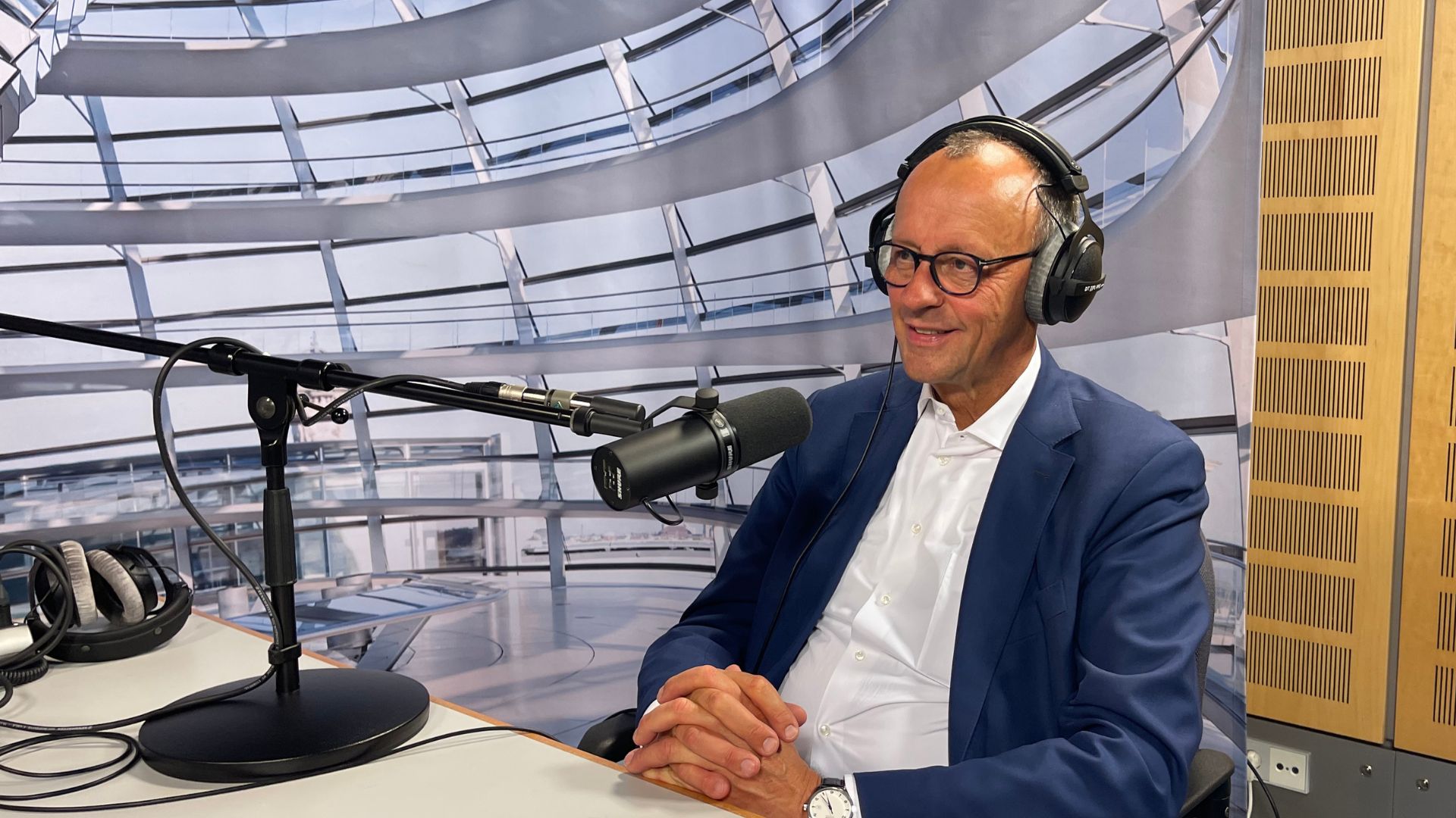 Ein Mann mit kurzen grauen Haaren, dunkler Brille und blauem Sakko spricht in ein Mikrofon. Er trägt große Kopfhörer.