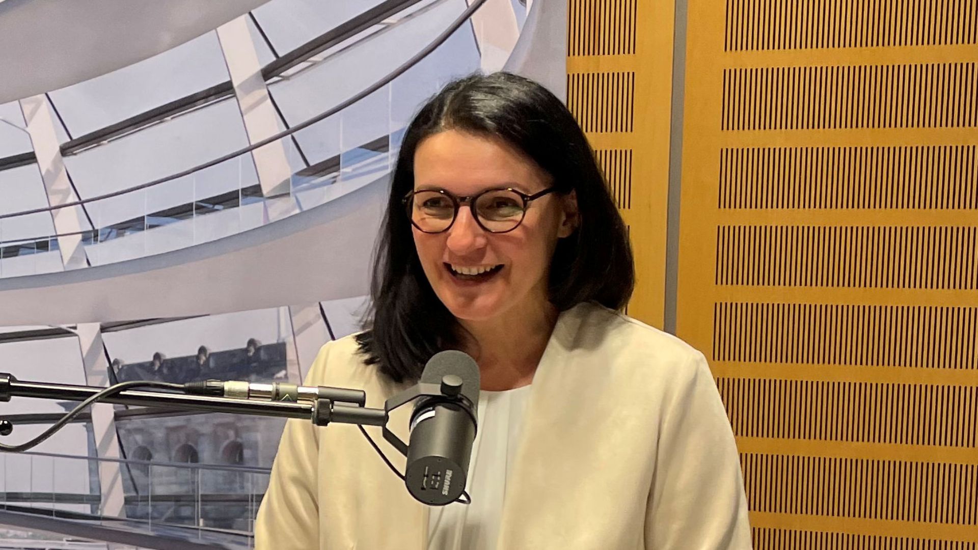 Eine Frau mit langen braunen Haaren und einer dunklen Brille spricht, in einen beigen Blazer gekleidet, in ein Mikrofon.