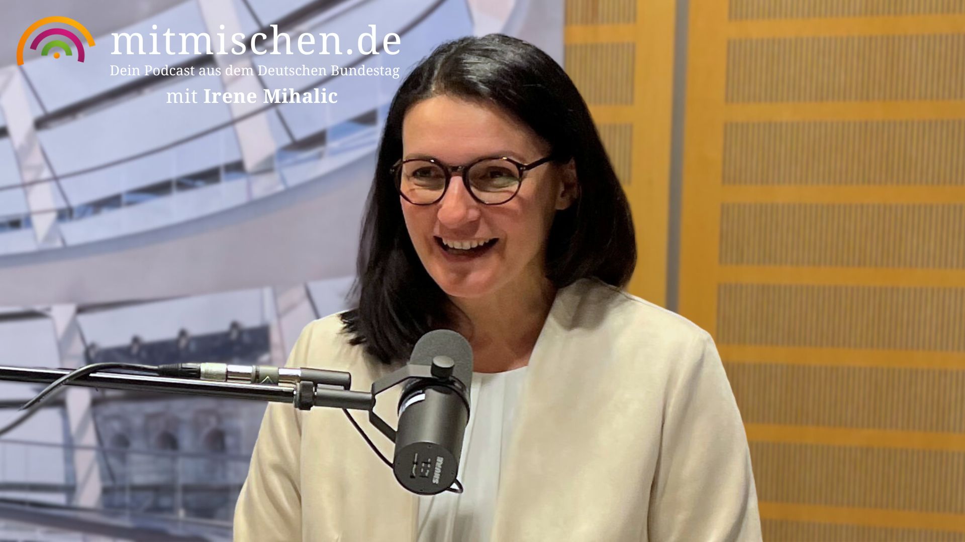 Eine Frau mit langen braunen Haaren und einer dunklen Brille spricht, in einen beigen Blazer gekleidet, in ein Mikrofon.