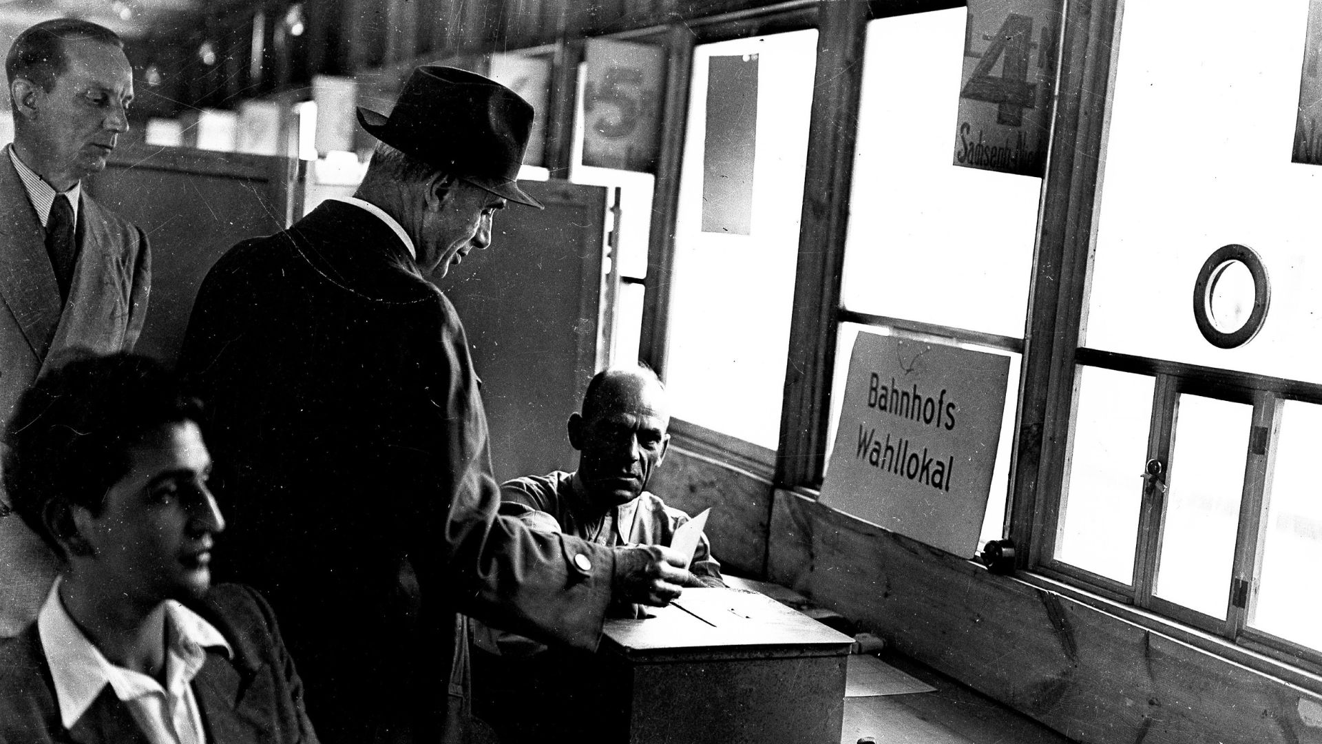 Ein Schwarz-Weiß-Foto auf dem vier Personen zu sehen sind. Ein Mann in Anzug und Hut wirft einen Stimmzettel in eine Box. Ihm gegenüber hängt ein Schild mit der Aufschrift "Bahnhofs Wahllokal"