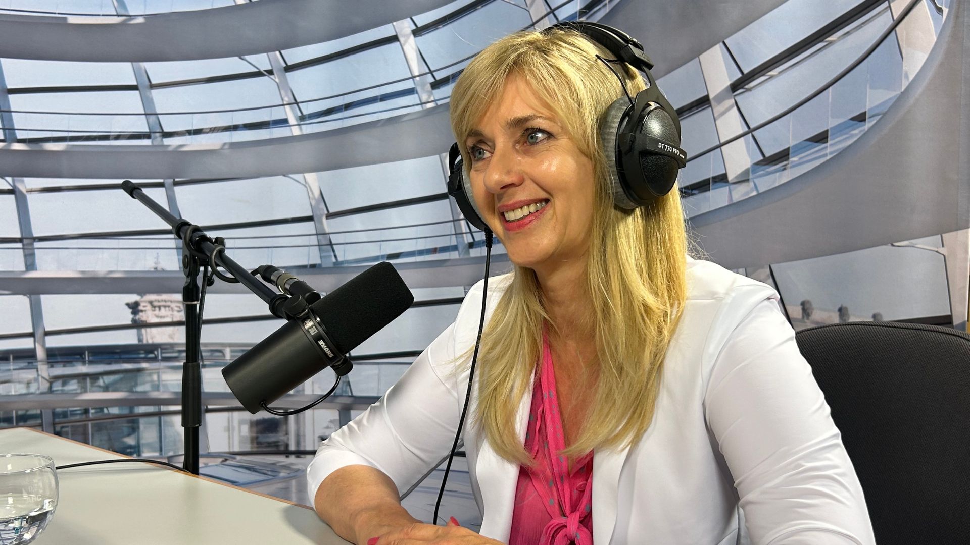 Eine Frau mit längeren blonden Haaren sitzt in einem Studio am Mikrofon. Sie trägt ein Pinkes Oberteil und einen weißen Blazer.